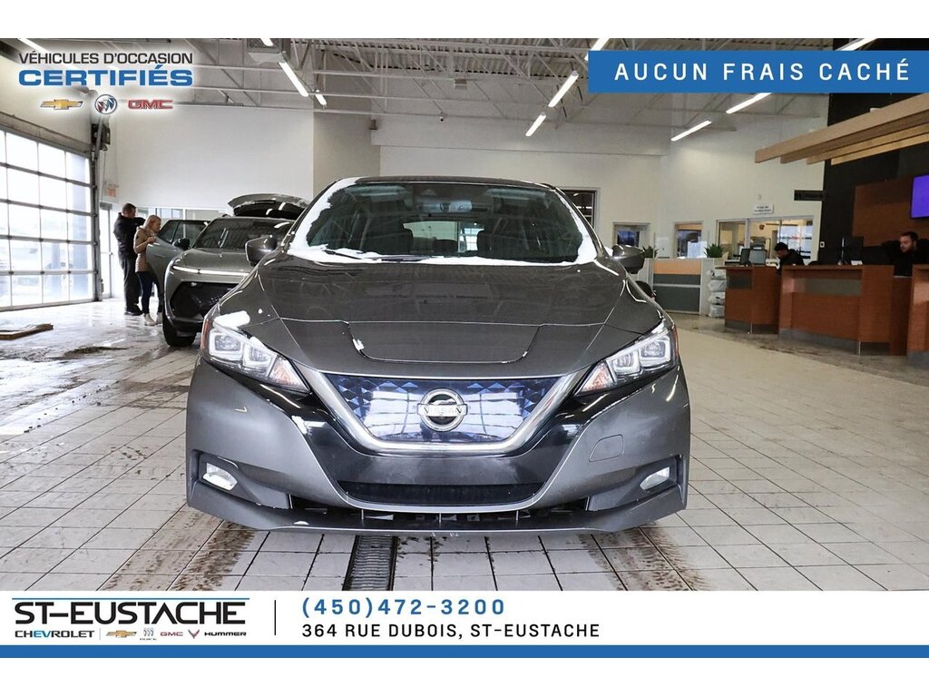 A Nissan Leaf electric vehicle which is dark grey in color is parked indoors at a car dealership showroom. The vehicle is clean and prominently displayed facing forward. In the background, dealership staff and customers are visible near other cars. The dealership branding for Chevrolet, Buick, GMC, and Hummer is visible at the top and bottom of the image, along with contact information for St-Eustache Chevrolet in Quebec, Canada. A banner in French at the top right reads "AUCUN FRAIS CACHÉ" (No Hidden Fees).