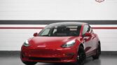 A red Tesla Model 3 electric vehicle parked indoors at Tabangi Motors dealership in Mississauga, Ontario. The car is facing forward with headlights on, and the dealership's branding and address are visible on the wall behind it.