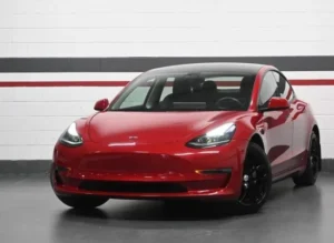 A red Tesla Model 3 electric vehicle parked indoors at Tabangi Motors dealership in Mississauga, Ontario. The car is facing forward with headlights on, and the dealership's branding and address are visible on the wall behind it.