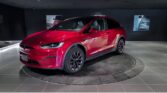 A red Tesla Model X electric SUV displayed on a rotating platform inside a modern showroom at Land Rover Richmond, located at 5680 Parkwood Crescent, Richmond, BC. The showroom features sleek black walls with illuminated vehicle posters. Branding and contact information for Range Rover Approved is shown at the bottom.