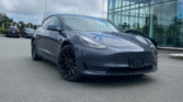 Front view of a dark gray Tesla Model 3 parked outdoors in a car lot under a cloudy sky."**