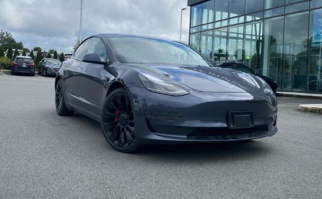 Front view of a dark gray Tesla Model 3 parked outdoors in a car lot under a cloudy sky."**